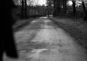 Black and white image photograph of a road with an out of focus foreground and a in focus background, nearing sunset